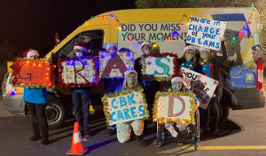 CBK students and staff in holiday attire hold signs with encouraging messages in front of the CBK van decorated with holiday lights.
